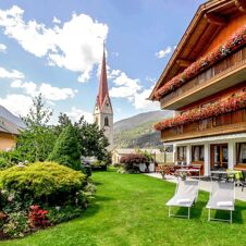 Hage med solstoler og balkongblomster p&aring; Hotel Waldheim foran en fjellkulisse og kirket&aring;rnet under en bl&aring; himmel.
