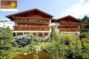 Fasaden p&aring; Hotel Waldheim viser et tradisjonelt trehus med balkonger og blomster, omgitt av en blomstrende hage med en dam.