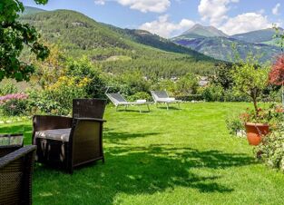 Outdoor garden area at Hotel Waldheim with grass, wicker chairs, sun loungers, flowering bushes, and forested mountains backdrop