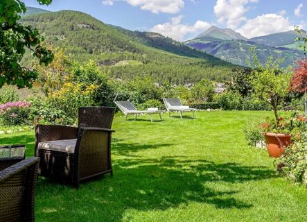 Outdoor garden area at Hotel Waldheim with grass, wicker chairs, sun loungers, flowering bushes, and forested mountains backdrop
