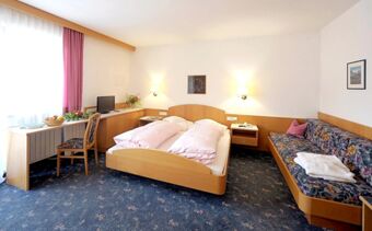 Hotel Waldheim room with double bed, floral carpet, wooden furniture, desk with chair, TV, and floral sofa with folded towels