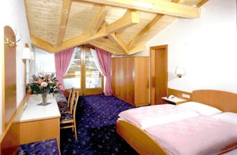 Bedroom with wooden ceiling beams, double bed, desk with chairs, wardrobe, balcony doors and pink curtains at Hotel Waldheim