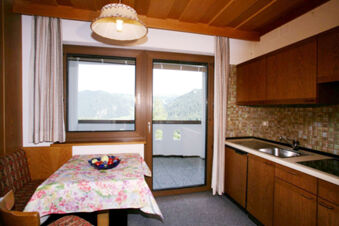 Small kitchen area with wooden cabinets, sink, stove, floral tablecloth on dining table, window, and door at Hotel Gasthof Borest & Residence Riposo