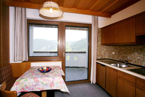 Small kitchen area with wooden cabinets, sink, stove, floral tablecloth on dining table, window, and door at Hotel Gasthof Borest & Residence Riposo