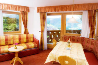 Living room with patterned orange and yellow sofa, wooden table with flower vase, and large windows at Hotel Gasthof Borest & Residence Riposo