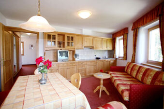 Living room with wooden kitchen cabinets, a vintage TV, red patterned sofa and floral tablecloth at Hotel Gasthof Borest & Residence Riposo