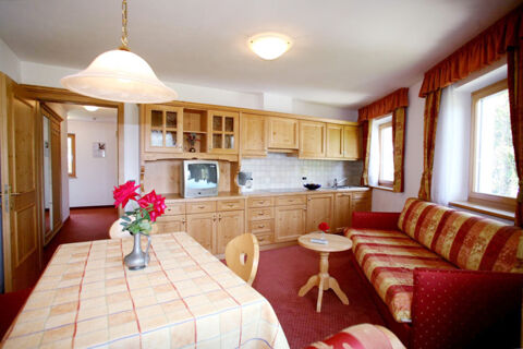Living room with wooden kitchen cabinets, a vintage TV, red patterned sofa and floral tablecloth at Hotel Gasthof Borest & Residence Riposo