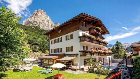Four-story building with balconies and flower boxes in front of a mountain, lawn with umbrella and playground at Hotel Maria