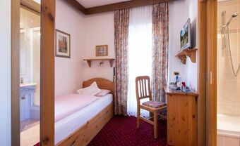 Single bed with pink bedding next to a window with floral curtains, wooden desk and chair, and visible bathroom in Hotel Maria