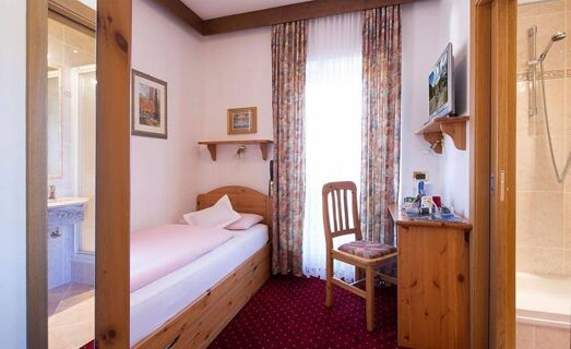 Single bed with pink bedding next to a window with floral curtains, wooden desk and chair, and visible bathroom in Hotel Maria