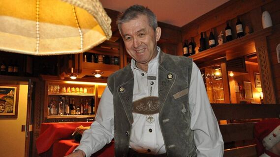 Homme en tenue traditionnelle souriant dans un bar confortable au d&eacute;cor de bois et de bouteilles de vin &agrave; l'h&ocirc;tel Maria.