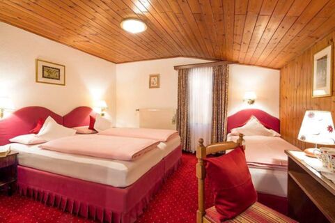 Hotel Maria guest room with two pink beds, red carpet, wooden ceiling, window with curtains, and bedside lamps