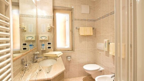 Bathroom at Hotel Maria with beige tiles, a sink with a granite countertop, wall-mounted toilet, and glass shower door visible