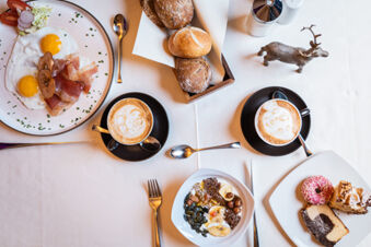 Breakfast spread at Hotel Fanes with fried eggs, bacon, bread rolls, cappuccinos, cereal bowl, and assorted cakes on a table