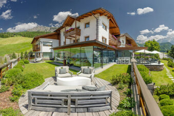 Outdoor seating area with cushions and glass-walled restaurant at Hotel Fanes surrounded by green lawns and mountains