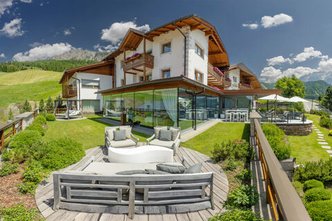 Outdoor seating area with cushions and glass-walled restaurant at Hotel Fanes surrounded by green lawns and mountains