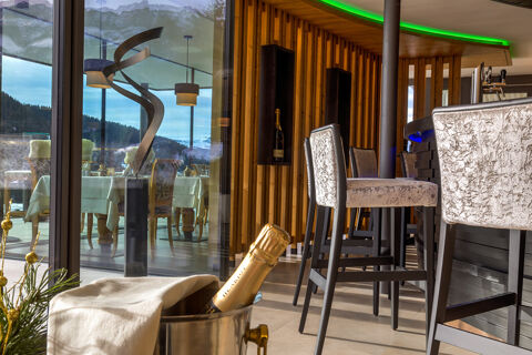 Interior view of Hotel Fanes bar area with silver textured chairs, wooden wall, champagne bottle in an ice bucket, and a reflection of dining tables outside