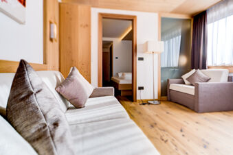 Living area in Hotel Fanes with beige sofas, gray cushions, wooden floors, a standing lamp, and a view into a bedroom with a bed