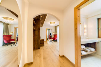Spacious hotel suite at Hotel Fanes with wooden floors, arches, red armchair, chandelier, and a bathroom with towels on shelves