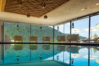 Indoor pool with woven chairs and hanging lights overlooking forested hills at Hotel Fanes through large glass windows