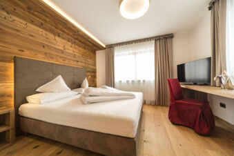 Hotel Fanes room with a large bed, wooden accent wall, red chair, desk with TV, and curtained window lighting the space