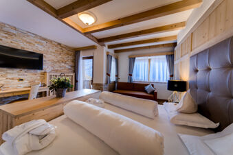 Hotel Fanes room with a bed, rolled white bedding, brown leather headboard, sofa, wooden desk, TV, and flower arrangement