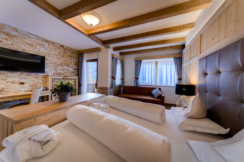Hotel Fanes room with a bed, rolled white bedding, brown leather headboard, sofa, wooden desk, TV, and flower arrangement