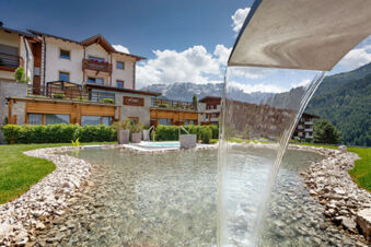 Waterfalls into a clear shallow pool surrounded by stones with green lawn and Hotel Fanes building and mountains in the background