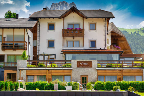 Hotel Fanes building with multiple balconies, glass windows, greenery, and mountain peaks under a blue sky background