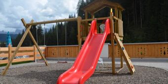 Utomhuslekplats med r&ouml;d rutschkana, gungor och kl&auml;tterv&auml;gg framf&ouml;r ett skogskl&auml;dd bergspanorama vid Hotel Schwarzenbach.