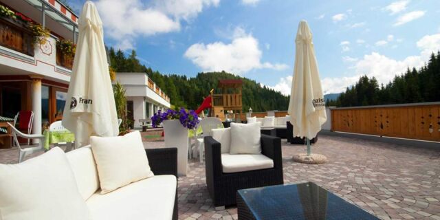 Terrasse med behagelige hvide siddepladser, parasoller og en legeplads med skoven som baggrund p&aring; Hotel Schwarzenbach.