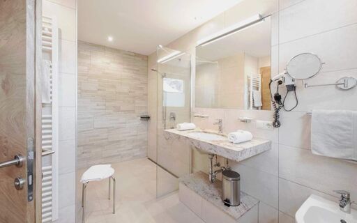 Bright bathroom in Hotel Theiner with a large mirror, marble sink, walk-in shower, stool, and hairdryer on the wall