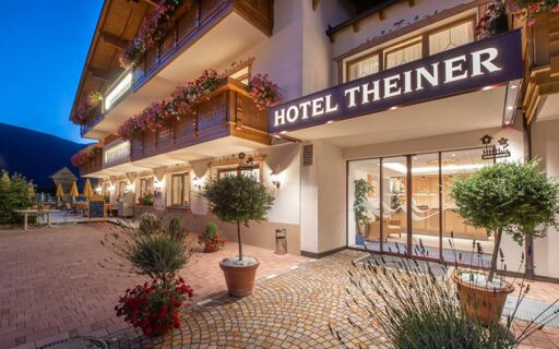 Hotel Theiner entrance at dusk with lit sign, wooden balconies adorned with flowers, and outdoor seating area with umbrellas