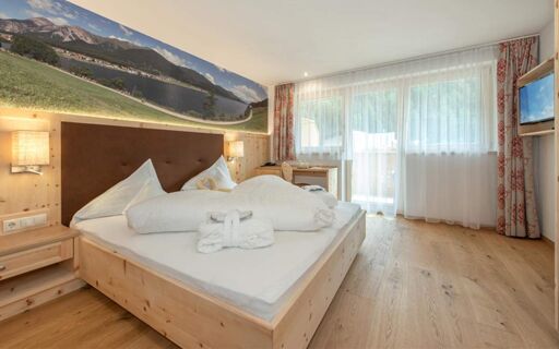 Hotel Theiner bedroom with wooden bed and bedside tables, mounted TV, sheer curtains, and mountain landscape wallpaper above headboard
