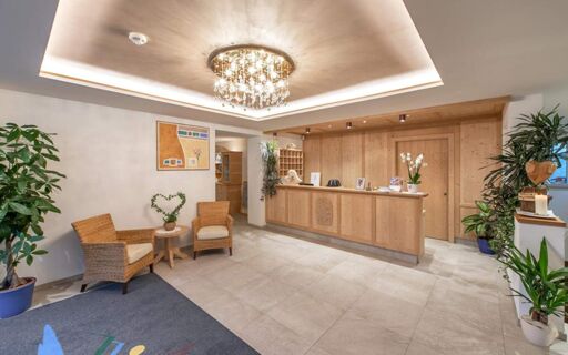 Reception area of Hotel Theiner with wooden front desk, crystal chandelier, two wicker chairs, indoor plants, and wall paintings