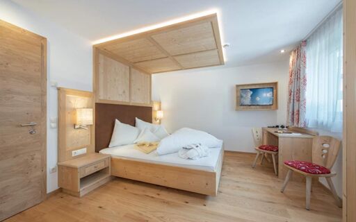 Hotel Theiner bedroom with wooden bed frame and headboard, white bedding, two chairs, desk, wall-mounted TV, and window with curtains