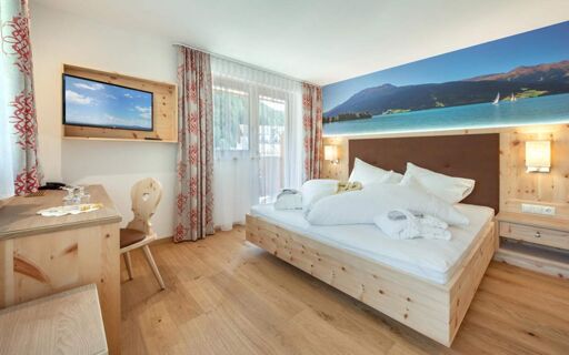 Hotel Theiner room with wooden furniture, double bed with white linens, wall-mounted TV, and large scenic wall mural of mountains and lake