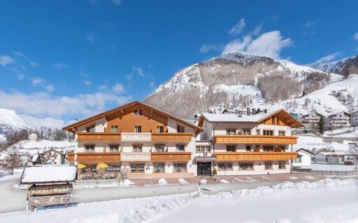 Het besneeuwde buitenaanzicht van Hotel Theiner toont een traditioneel Alpenhotel met houten balustrades tegen een bergachtige achtergrond.