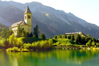 A lake reflects trees, a church with a tall steeple, and Hotel Alpenhof near the base of a large mountain range