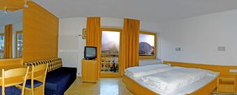 Hotel Alpenhof room with double bed, wooden furniture, TV, blue sofa, yellow curtains, and mountain view through windows