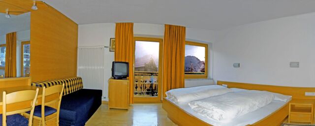 Hotel Alpenhof room with double bed, wooden furniture, TV, blue sofa, yellow curtains, and mountain view through windows