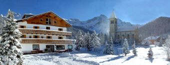 Hotel Alpenhof building covered with snow next to a church with mountains and snow-covered trees under a clear blue sky