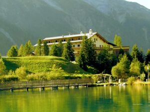 Hotel Alpenhof surrounded by trees near a wooden bridge over calm water with mountains in the background
