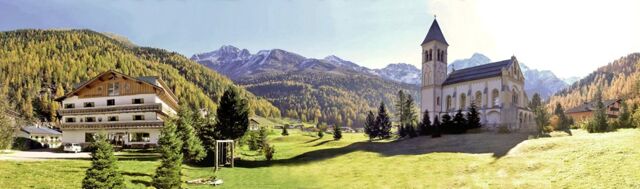 Hotel Alpenhof surrounded by green grass, evergreen trees, a large white church with a tower, and mountains in the background