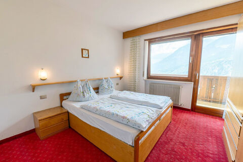 Double bed with floral bedding, wooden furniture, red carpet, wall lights, and mountain view window at Residence Haus am Berg