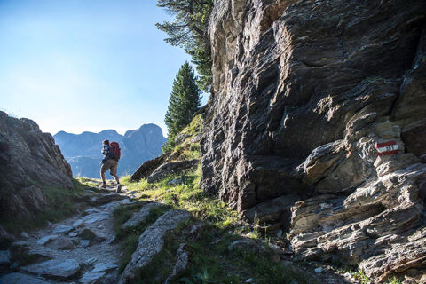A hiker with a red backpack walking on a rocky mountain path near a cliff with a trail marker 151 at Residence Haus am Berg