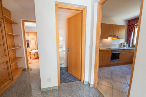 Interior view of a hallway with wooden shelves, an open bathroom door, kitchen with cabinets, and text Residence Haus am Berg