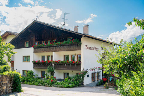 Residence Haus am Berg is a white multi-story building with wooden balconies adorned with red flowers and surrounded by green plants under a blue sky