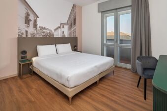 King-size bed with white linens in a room featuring a wall mural, side tables, gray curtains, and a balcony door at B&B Hotel Treviso Al Fogher