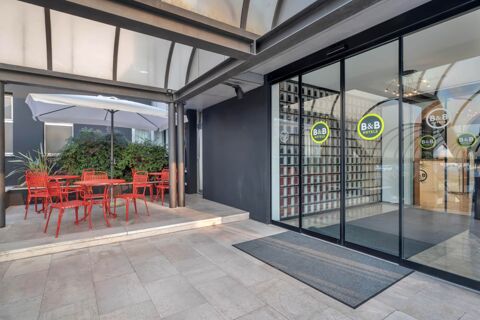 Entrance of B&B Hotel Treviso Al Fogher showing glass sliding doors with logos and outdoor seating area with red chairs and tables under white umbrella